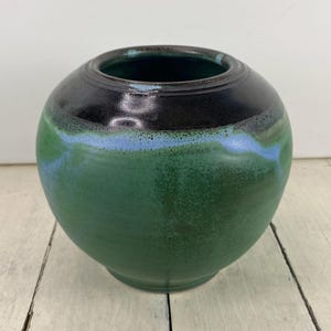 May include: A round ceramic vase with a dark green base, transitioning to a light blue band, and a black rim. The vase has a smooth, glossy finish and a wide opening. The vase is sitting on a white wooden surface.