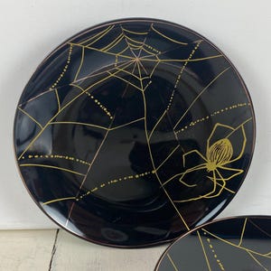 May include: Black ceramic plate with a gold spiderweb design and a gold spider. The plate has a brown rim. The plate is a Halloween-themed decorative item.