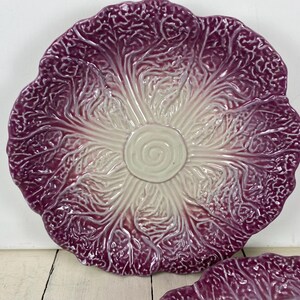 May include: A round ceramic plate shaped like a head of radicchio or red cabbage. The surface features a textured, embossed leaf pattern with a deep purple color at the edges that fades into a creamy white center with a spiral design.