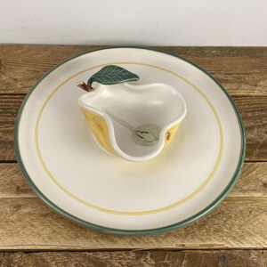 May include: A ceramic serving set consisting of a large round off-white plate with a thin yellow inner ring and a dark green outer rim, paired with a pear-shaped bowl featuring a yellow exterior, a white interior, and a green leaf handle.