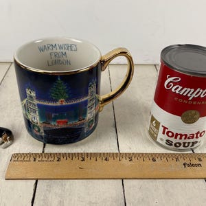 K&ouml;nnte beinhalten: Eine blaue Keramiktasse mit goldenem Henkel und Rand, die eine Londoner Szene und den Text "Warm Wishes From London" zeigt. Eine Dose Campbell's Tomatensuppe und eine Corgi-Figur sind ebenfalls vorhanden.