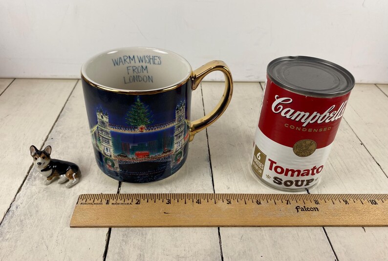 K&ouml;nnte beinhalten: Eine blaue Keramiktasse mit goldenem Henkel und Rand, die eine Londoner Szene und den Text "Warm Wishes From London" zeigt. Eine Dose Campbell's Tomatensuppe und eine Corgi-Figur sind ebenfalls vorhanden.