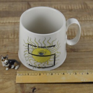 Pode incluir: Caneca de cer&acirc;mica branca com um desenho de sol amarelo e uma letra preta "S" no centro. O sol tem um olho azul no centro.