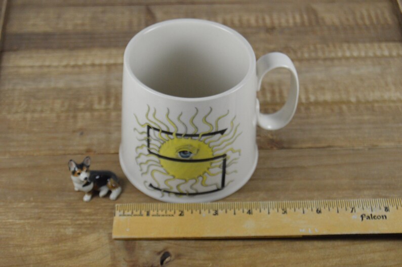 Pode incluir: Caneca de cer&acirc;mica branca com um desenho de sol amarelo e uma letra preta "S" no centro. O sol tem um olho azul no centro.