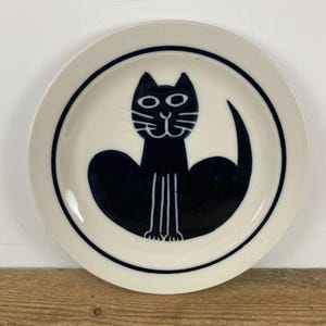 May include: A white ceramic plate with a black cat illustration. The cat is depicted in a minimalist style with a black body, tail, and facial features. A black line borders the plate's edge.