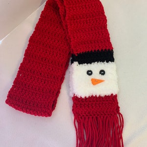 May include: Red crocheted scarf with a snowman face on one end. The snowman has black eyes, an orange nose, and a white fuzzy face.