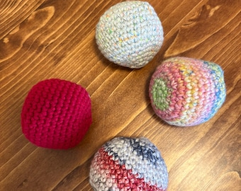 May include: Four handmade crochet balls in various colors rest on a wooden surface. One is bright red, another is a mix of pastel colors, and a third is a mix of red, white, and blue. The fourth ball is light blue with colorful flecks.