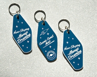 May include: Three dark blue motel-style keychains with silver key rings. Two keychains read "Never Sleeping Always Dreaming" with star graphics. The third keychain says "Cosmic Comet Motel" with a rocket ship graphic.