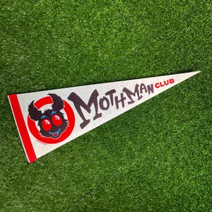 May include: A light gray pennant with the words "Mothman Club" in black and red. The pennant features a cartoon moth creature with red eyes and a blue smile inside a red circle. The pennant has a red border.