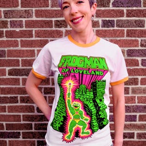 May include: White ringer t-shirt with an orange collar and sleeve trim. The shirt features a graphic of a green frog character holding a glowing staff, with the words "FROGMAM OF LOVELAND" in green and pink.