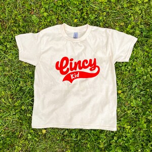 Puede incluir: Camiseta de color crema con las palabras "Cincy Kid" en letras rojas. El texto está en una fuente de estilo béisbol. La camiseta está hecha de un material suave y es adecuada para niños.