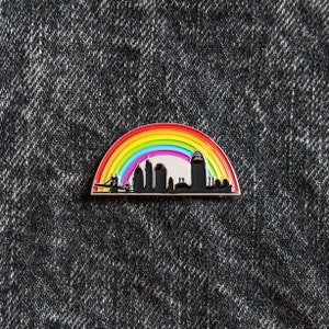 Op de afbeelding: Een regenboog-emaille pin met een silhouet van een stadsbeeld met een brug en gebouwen. De pin is op een donkerblauwe denim achtergrond.