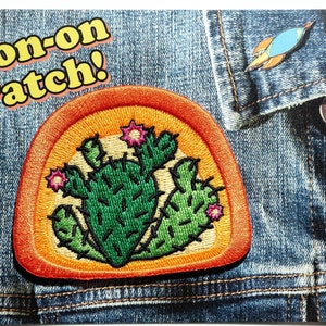 May include: Iron-on patch with a cactus design. The patch is orange with a yellow center and features a green cactus with pink flowers. The text "Iron-on Patch!" is in yellow letters on a pink background.