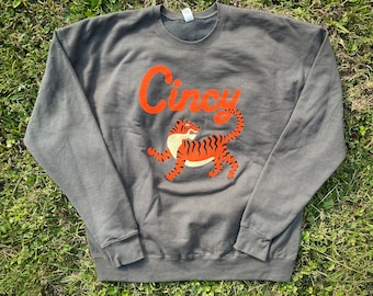 May include: A gray sweatshirt with the word "Cincy" in orange script above a cartoon tiger. The tiger is orange with black stripes and is facing right. The sweatshirt is laying on green grass.