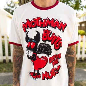May include: White ringer t-shirt with a red collar and cuffs. The shirt features a cartoon Mothman character with a red heart-shaped butt and the text "Mothman Butts Drive Me Nuts!" in red.