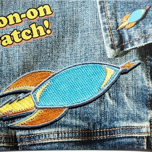 May include: A denim jacket features a blue, orange, and yellow embroidered rocket iron-on patch. The text "Iron-on Patch!" is above the patch. A smaller rocket pin is also visible.