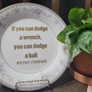 May include: A decorative white plate with a floral border and gold trim, displaying the quote "If you can dodge a wrench, you can dodge a ball" attributed to Patches O'Houlihan. The plate is on a metal stand, next to a potted plant.