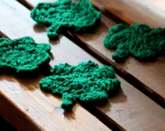 May include: Four green crocheted shamrock-shaped coasters on a wooden surface.