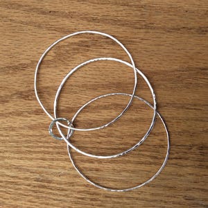 May include: Three silver bangles stacked on top of each other on a wooden surface.