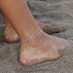 Może przedstawiać: Boso stopy i dolne nogi osoby spoczywają na piaszczystej plaży. Osoba nosi srebrną bransoletkę na kostkę z zawieszką w kształcie nieskończoności i małą turkusową koralikiem.