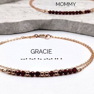 以下が含まれることがあります： 赤い宝石のビーズが付いた、繊細なゴールドチェーンのブレスレットが2つ。上のブレスレットには「MOMMY」と書いてあり、下のブレスレットには「GRACIE」と書いてあり、その下に点字が書かれています。