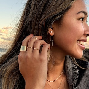 May include: A woman wearing gold jewelry, including rings, earrings, and necklaces. She is also wearing a gray and black patterned shawl and a blue sweater. The background shows a beach and the ocean.