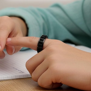 Può includere: Un anello nero antistress è sul dito di una persona. L'anello ha un design strutturato. L'immagine include il testo "Keep Hands Busy, Minds Sharp". La persona indossa una camicia azzurra e siede a una scrivania di legno.
