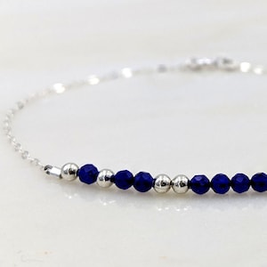 May include: A fine silver bracelet with a string of dark blue, faceted beads and silver beads. The bracelet has a minimalist design, suitable for any occasion. The name "Cathie" is written below the bracelet in black text, along with a morse code sequence.
