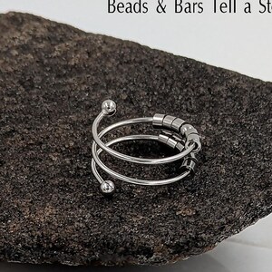 Puede incluir: Un anillo plateado en forma de espiral con pequeñas cuentas redondas y el texto "Spin Gently Beads & Bars Tell a Story". El anillo se muestra sobre una superficie oscura y texturizada. También es visible la palabra "Mommy".