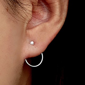 Può includere: Primo piano di un orecchio con due orecchini d'argento. Uno è un piccolo bottone, l'altro è un orecchino a cerchio aperto e curvo. Gli orecchini sono appoggiati sulla pelle dell'orecchio e sull'area circostante.