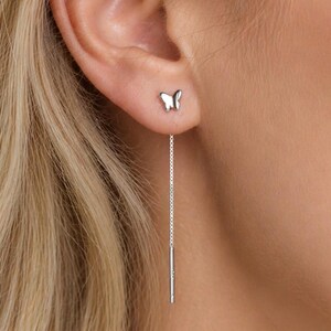 May include: A close-up of a woman's ear adorned with two silver earrings. One is a small butterfly stud, and the other is a long, dangling chain with a small bar at the end. The earrings are set against the backdrop of the woman's blonde hair.