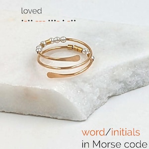 Puede incluir: Un anillo en espiral de color dorado con cuentas plateadas. El anillo está inscrito con la palabra "loved" en código Morse. También se ve el texto "word/initials in Morse code".