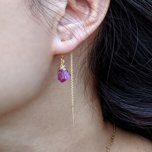 Puede incluir: Un par de pendientes de cadena dorada con una piedra preciosa rosa que cuelga de cada cadena. Los pendientes los lleva una persona con el pelo largo y oscuro.