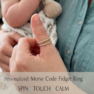Puede incluir: Un anillo plateado con dos bandas, adornado con pequeñas cuentas doradas, se lleva en un pulgar. El anillo es un anillo fidget de código Morse. El texto en la imagen dice: "Personalized Morse Code Fidget Ring" y "SPIN TOUCH CALM."
