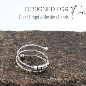 May include: A silver fidget ring with a spiral design and small, rolling beads. The ring is displayed on a textured, gray stone. Text on the image reads "DESIGNED FOR Focus Quiet Fidget & Restless Hands."