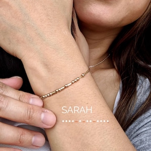 Peut inclure: Un bracelet en perles dorées avec un pendentif en forme de barre gravé du nom "SARAH".
