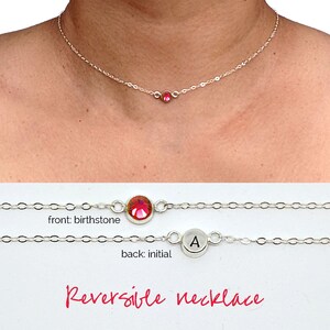 May include: A silver chain necklace with a red birthstone on one side and a silver charm with the letter "A" on the other side. The necklace is reversible and says "Reversible necklace" on the white background.