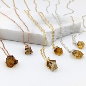 May include: Five gold, rose gold, and silver necklaces with raw citrine crystal pendants. The necklaces are displayed on a white marble surface.