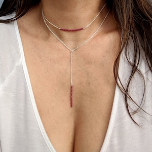 Peut inclure: Un collier en argent avec un long pendentif fin composé de perles rouges. Le collier est superposé à une chaîne plus courte et plus fine de perles rouges.