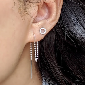 Op de afbeelding: Een paar zilveren oorbellen met een delicate ketting die aan één oorbel hangt. De andere oorbel is een kleine cirkel met heldere stenen.