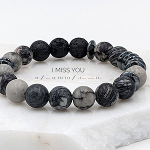May include: A black and grey beaded bracelet with a lava rock bead and metal accents. The bracelet has the text "I MISS YOU" and braille dots that read "I MISS YOU".