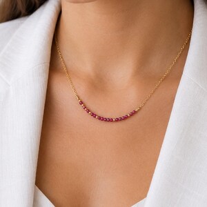 May include: A gold chain necklace featuring a curved center section adorned with a row of small, round, deep pink beads interspersed with tiny gold spheres, resting against a white collared garment.