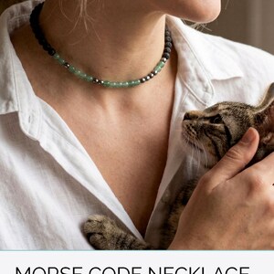 May include: A beaded necklace with black, green, and silver beads. The necklace is around a person's neck. The image includes the text "MORSE CODE NECKLACE in your Cat's Name & Birthstone."