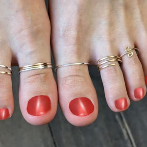 May include: Close-up of two feet with red nail polish, each toe adorned with delicate toe rings. The rings vary in design, including simple bands and spiral styles, in gold and silver tones.