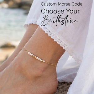 May include: A gold anklet featuring small white pearls and gold beads. The anklet is worn on an ankle, with the text "Custom Morse Code Choose Your Birthstone" in the background. The person is wearing a white dress.