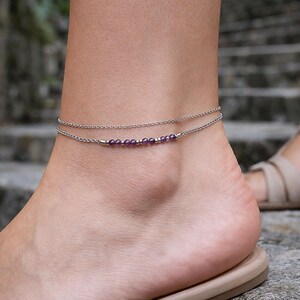 May include: A double-strand silver-toned anklet featuring a delicate chain design. The lower strand is accented with a series of small, round, purple amethyst beads interspersed with tiny metallic silver-toned spacer beads in a linear arrangement.