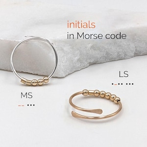 Puede incluir: Dos anillos, uno plateado y otro dorado, cada uno con pequeñas cuentas doradas. Los anillos se muestran sobre una superficie blanca. El texto dice "iniciales en código Morse" con las iniciales "MS" y "LS" en código Morse.