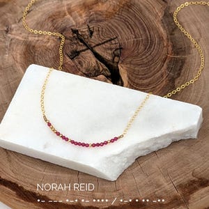 May include: A delicate gold chain necklace featuring a central row of small, vibrant pink beads interspersed with tiny gold spacers. The necklace is displayed on a white stone slab, with the text NORAH REID and Morse code printed below.