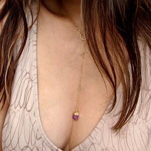 May include: A gold chain necklace with a small, pink crystal pendant. The necklace is worn by a person with long brown hair.