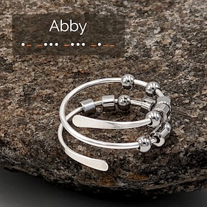 Puede incluir: Un anillo en espiral plateado con cuentas móviles y detalles cilíndricos. El anillo tiene el nombre "Abby" encima. El texto debajo dice: "This spiral style is trickier to adjust: pls indicate US ring size in Notes at checkout & I'll make it closer to that size."